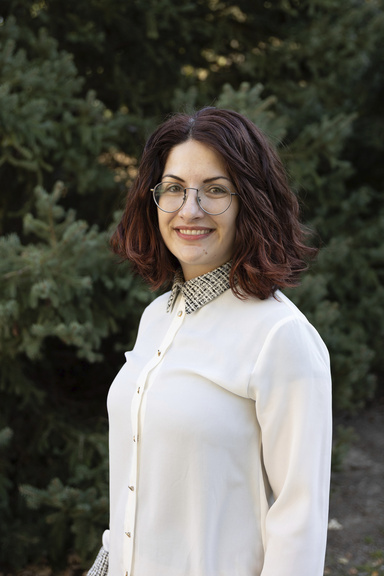 A smiling woman in a white blouse poses in front of some greenery.