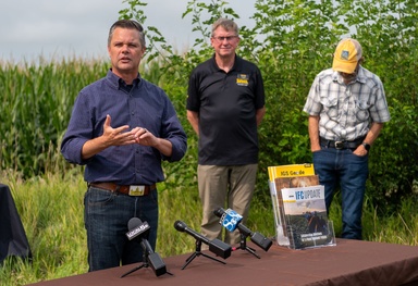 Zach Nunn stands behind a table with media microphones, speaking to the crowd, with a cornfield behind him and two men listening