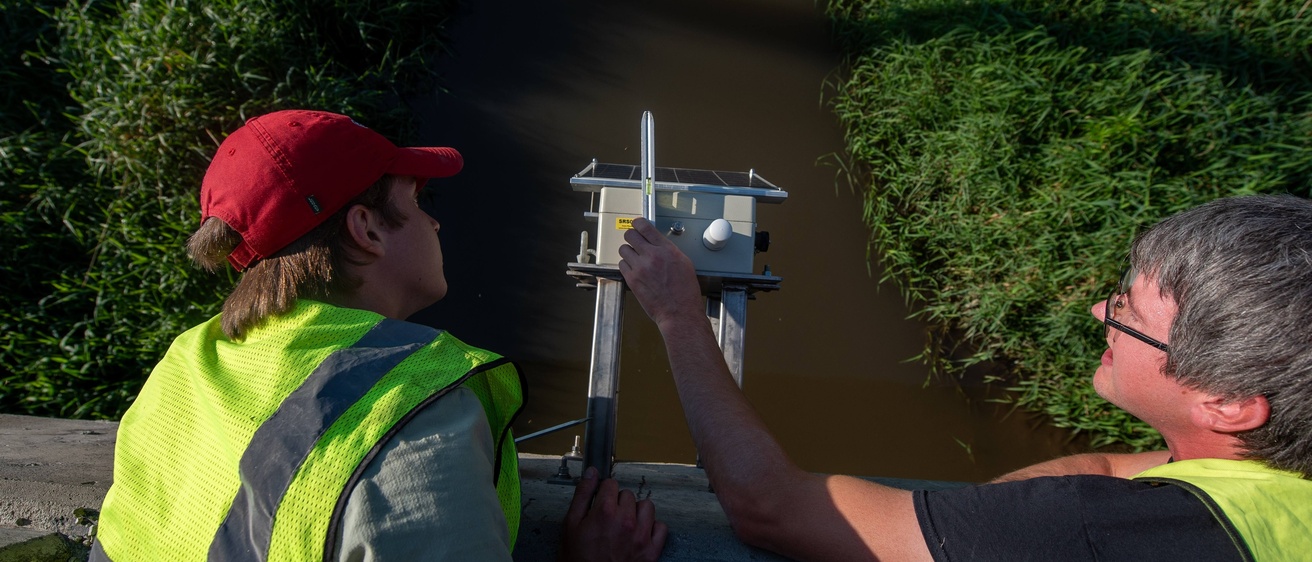IFC employees secure a bridge sensor on a concrete bridge over a stream