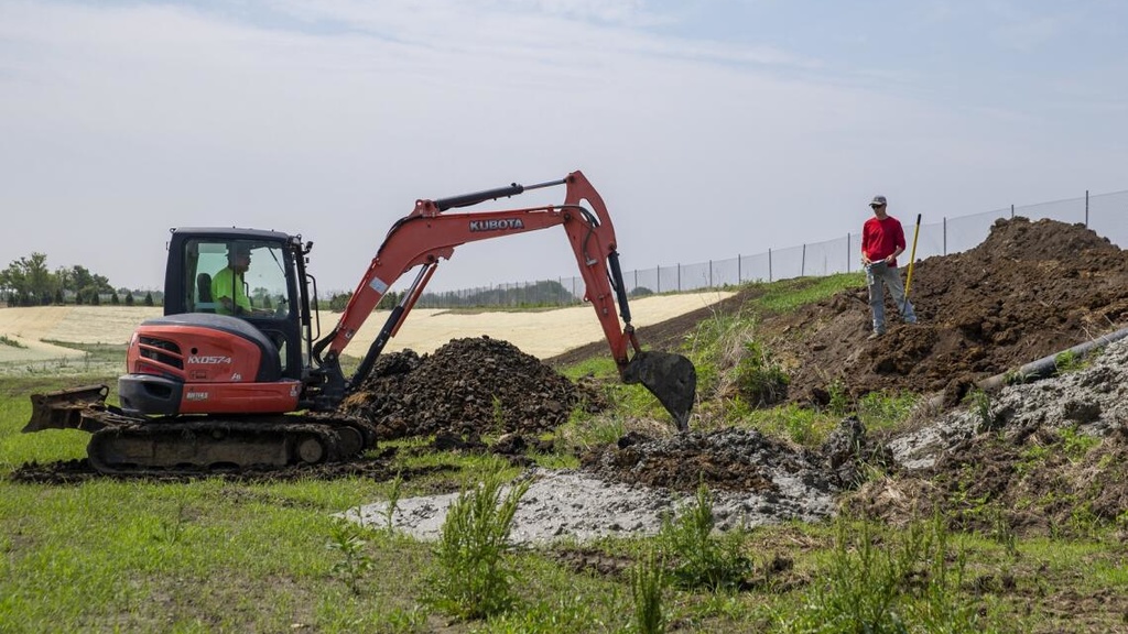 A person monitors a pipe as a construction vehicle digs out dirt around it 